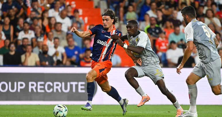 Soccer player in blue-and-orange striped jersey dribbles the ball as two gray-clad defenders close in on a green field with a cheering crowd behind.