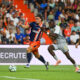 Soccer player in blue-and-orange striped jersey dribbles the ball as two gray-clad defenders close in on a green field with a cheering crowd behind.
