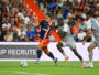 Soccer player in blue-and-orange striped jersey dribbles the ball as two gray-clad defenders close in on a green field with a cheering crowd behind.