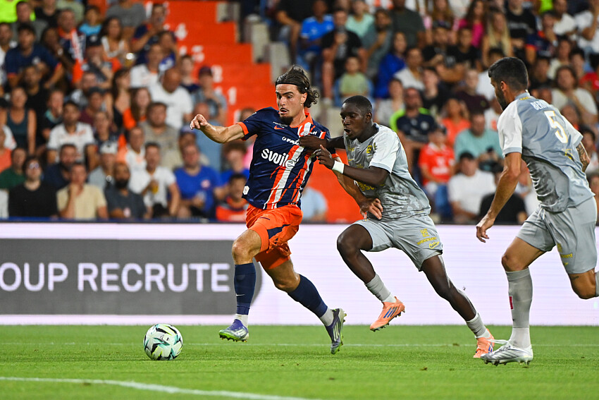 Pays | AllezPaillade.com Soccer player in blue-and-orange striped jersey dribbles the ball as two gray-clad defenders close in on a green field with a cheering crowd behind.