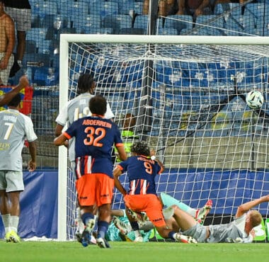 Soccer goal scene with players in blue-orange and gray jerseys as the ball hits the net; goalkeeper on the ground near the post while teammates react nearby.