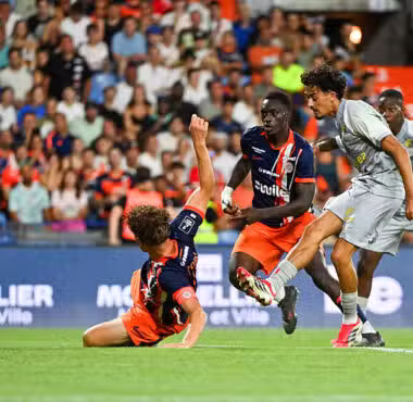Two soccer players contest the ball during a sliding tackle as a gray-uniform opponent approaches in a crowded stadium.
