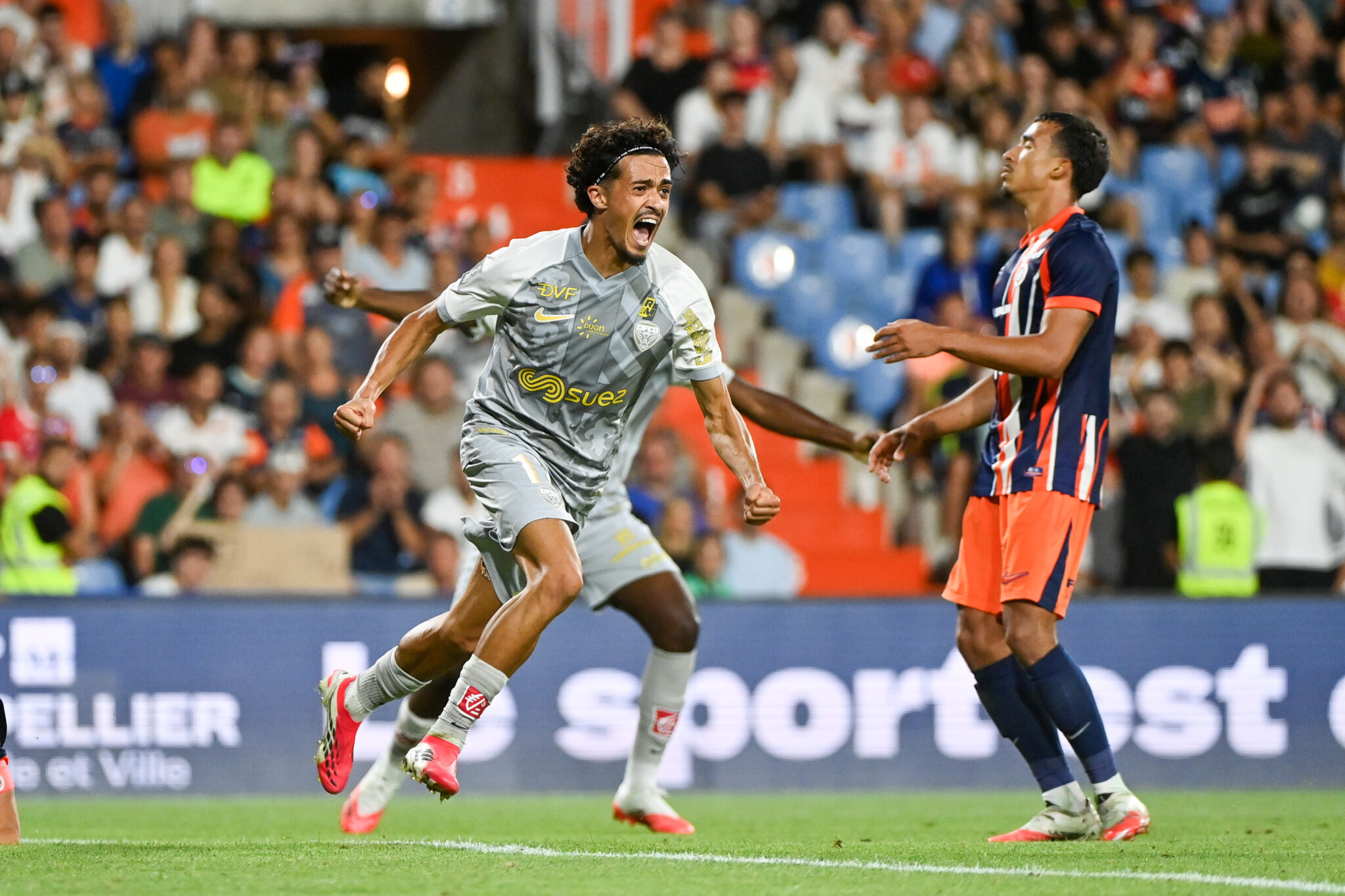 Montpellier Herault SC v Dijon FCO - Ligue 2 BKT 20262027 | AllezPaillade.com Soccer player in a gray kit sprints forward, shouting, as a defender in blue‑red stripes pursues amid a packed crowd.