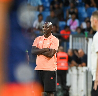 Male coach in a peach polo and black shorts stands with arms crossed on the sideline during a soccer match, crowd in the background.