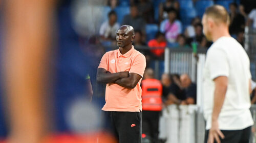 Male coach in a peach polo and black shorts stands with arms crossed on the sideline during a soccer match, crowd in the background.