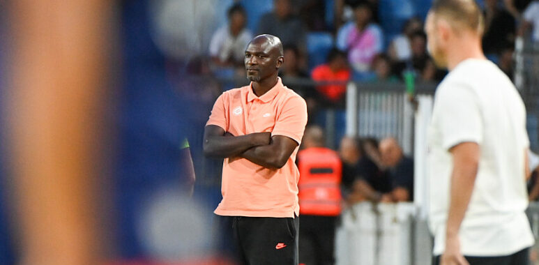 Male coach in a peach polo and black shorts stands with arms crossed on the sideline during a soccer match, crowd in the background.