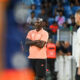 Male coach in a peach polo and black shorts stands with arms crossed on the sideline during a soccer match, crowd in the background.