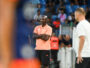 Male coach in a peach polo and black shorts stands with arms crossed on the sideline during a soccer match, crowd in the background.
