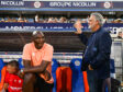 Two men by a soccer dugout: a dark-skinned man in a peach polo sits on orange-blue seats, while a gray-haired man in a navy blazer stands beside him with a railing touch; stands and spectators in the background.