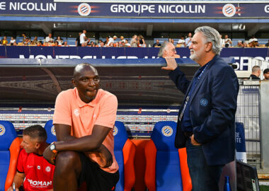 Two men by a soccer dugout: a dark-skinned man in a peach polo sits on orange-blue seats, while a gray-haired man in a navy blazer stands beside him with a railing touch; stands and spectators in the background.