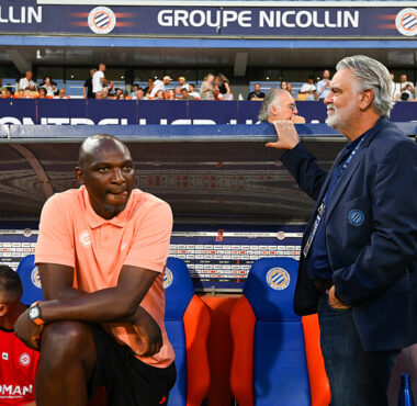 Two men by a soccer dugout: a dark-skinned man in a peach polo sits on orange-blue seats, while a gray-haired man in a navy blazer stands beside him with a railing touch; stands and spectators in the background.