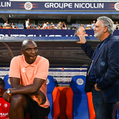 Two men by a soccer dugout: a dark-skinned man in a peach polo sits on orange-blue seats, while a gray-haired man in a navy blazer stands beside him with a railing touch; stands and spectators in the background.