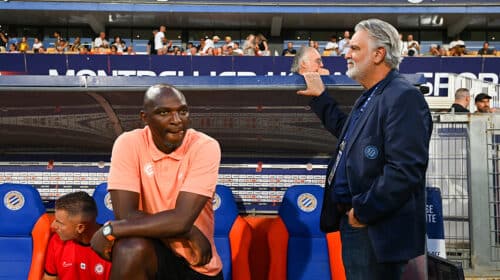 Two men by a soccer dugout: a dark-skinned man in a peach polo sits on orange-blue seats, while a gray-haired man in a navy blazer stands beside him with a railing touch; stands and spectators in the background.