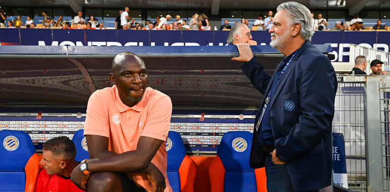 Two men by a soccer dugout: a dark-skinned man in a peach polo sits on orange-blue seats, while a gray-haired man in a navy blazer stands beside him with a railing touch; stands and spectators in the background.