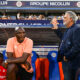 Two men by a soccer dugout: a dark-skinned man in a peach polo sits on orange-blue seats, while a gray-haired man in a navy blazer stands beside him with a railing touch; stands and spectators in the background.