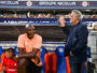 Two men by a soccer dugout: a dark-skinned man in a peach polo sits on orange-blue seats, while a gray-haired man in a navy blazer stands beside him with a railing touch; stands and spectators in the background.