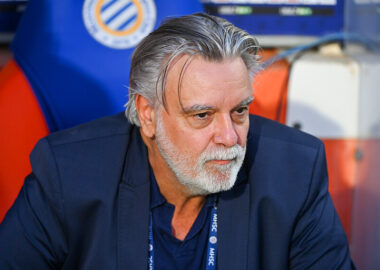 Older man with gray hair and beard in a navy blazer, seated in a sports dugout, looking thoughtful.