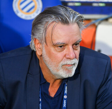 Older man with gray hair and beard in a navy blazer, seated in a sports dugout, looking thoughtful.