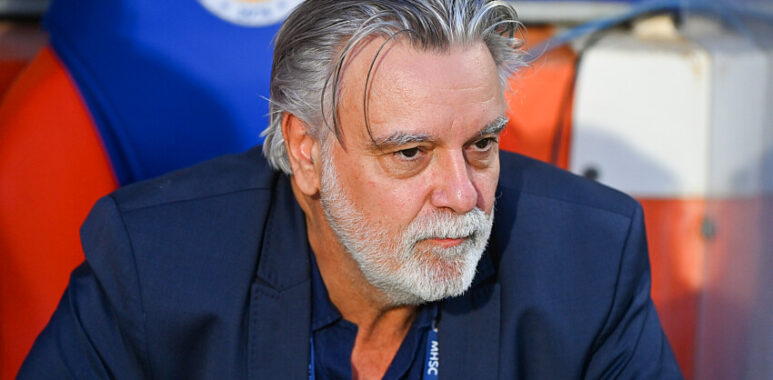 Nicollin | AllezPaillade.com Older man with gray hair and beard in a navy blazer, seated in a sports dugout, looking thoughtful.