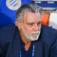 Older man with gray hair and beard in a navy blazer, seated in a sports dugout, looking thoughtful.