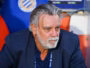 Older man with gray hair and beard in a navy blazer, seated in a sports dugout, looking thoughtful.