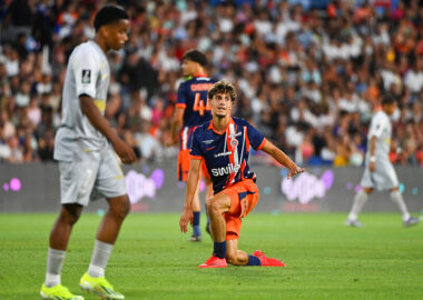 Soccer player in orange and navy kit kneels on the grass with one arm out, during a match in a packed stadium.