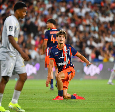 Soccer player in orange and navy kit kneels on the grass with one arm out, during a match in a packed stadium.