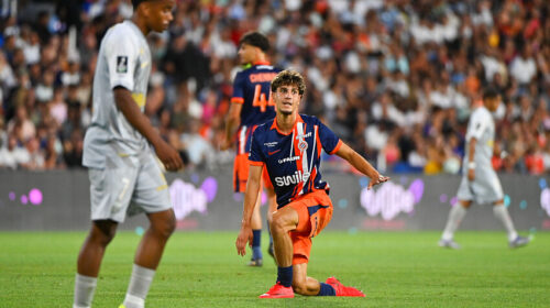 Soccer player in orange and navy kit kneels on the grass with one arm out, during a match in a packed stadium.