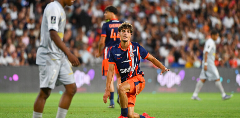 Noah Vidal-Cartoux | AllezPaillade.com Soccer player in orange and navy kit kneels on the grass with one arm out, during a match in a packed stadium.