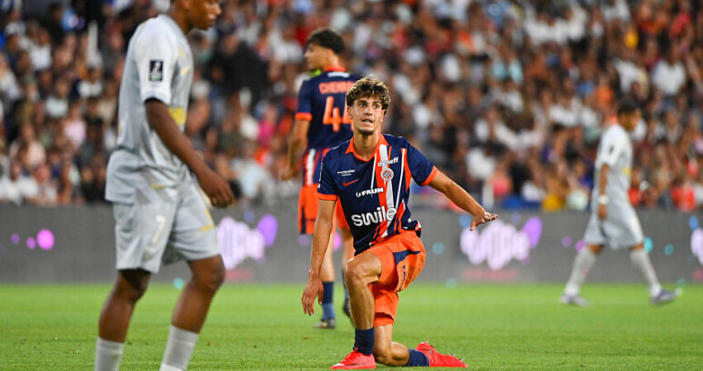 Soccer player in orange and navy kit kneels on the grass with one arm out, during a match in a packed stadium.