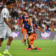 Soccer player in orange and navy kit kneels on the grass with one arm out, during a match in a packed stadium.