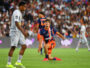 Soccer player in orange and navy kit kneels on the grass with one arm out, during a match in a packed stadium.