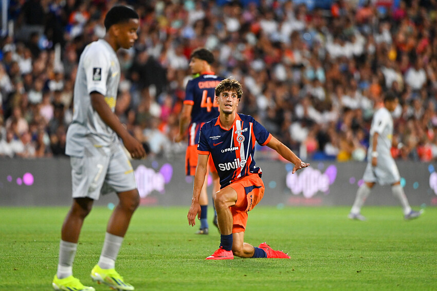 Noah Vidal-Cartoux | AllezPaillade.com Soccer player in orange and navy kit kneels on the grass with one arm out, during a match in a packed stadium.