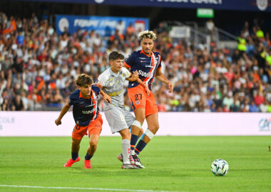 Soccer players in orange and navy jerseys contest the ball on a crowded stadium pitch; two players challenge a gray-uniformed opponent as the ball nears the sideline, with a full audience in the background.