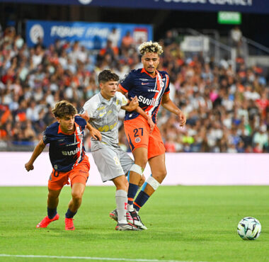Soccer players in orange and navy jerseys contest the ball on a crowded stadium pitch; two players challenge a gray-uniformed opponent as the ball nears the sideline, with a full audience in the background.