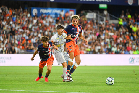 Soccer players in orange and navy jerseys contest the ball on a crowded stadium pitch; two players challenge a gray-uniformed opponent as the ball nears the sideline, with a full audience in the background.