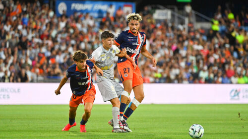 Soccer players in orange and navy jerseys contest the ball on a crowded stadium pitch; two players challenge a gray-uniformed opponent as the ball nears the sideline, with a full audience in the background.