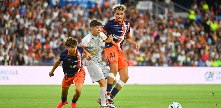 Soccer players in orange and navy jerseys contest the ball on a crowded stadium pitch; two players challenge a gray-uniformed opponent as the ball nears the sideline, with a full audience in the background.
