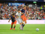 Soccer players in orange and navy jerseys contest the ball on a crowded stadium pitch; two players challenge a gray-uniformed opponent as the ball nears the sideline, with a full audience in the background.