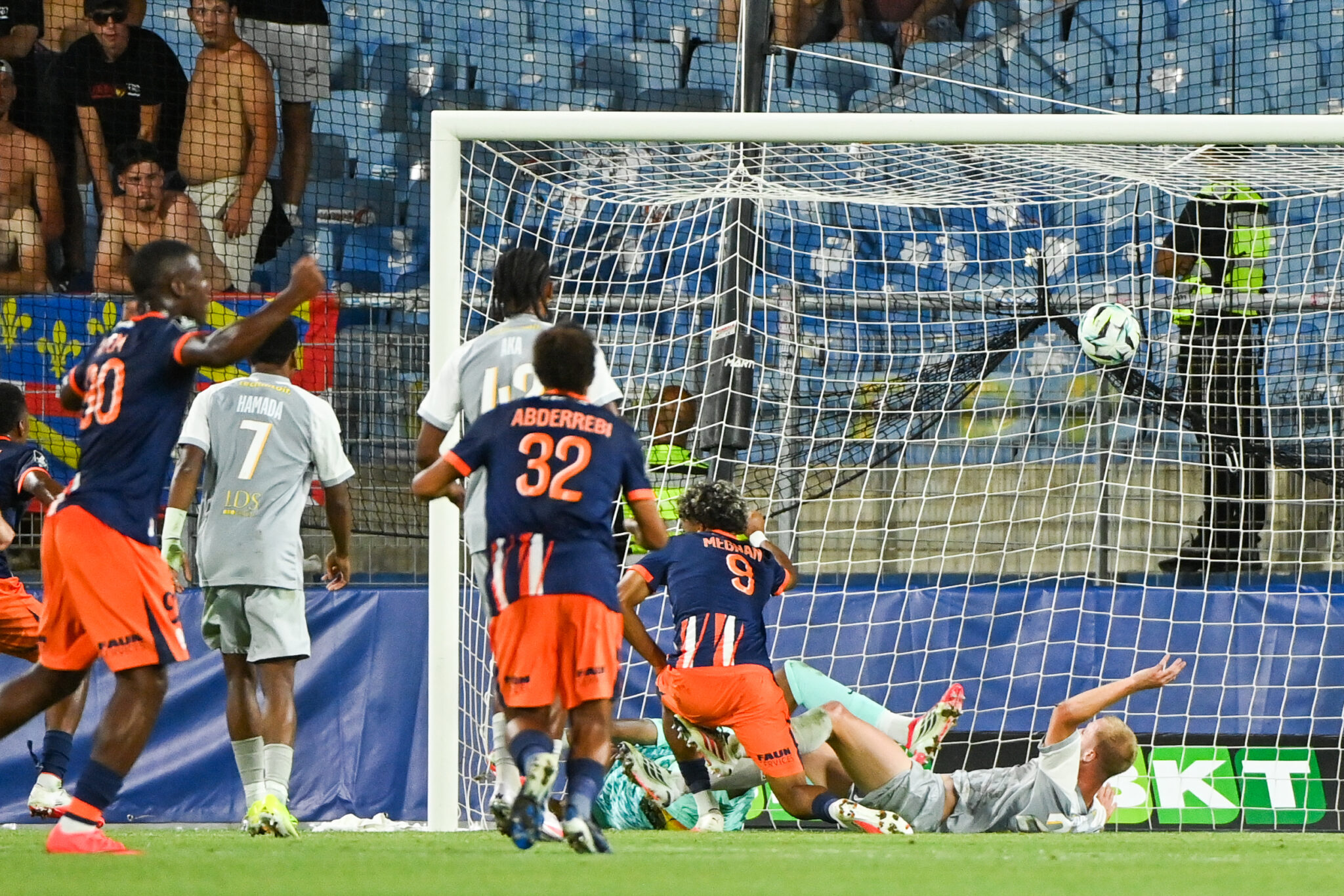 Montpellier Herault SC v Dijon FCO - Ligue 2 BKT 20262027 | AllezPaillade.com Soccer match scene: players in blue and orange chase the ball near the goal as the goalkeeper lies on the ground.
