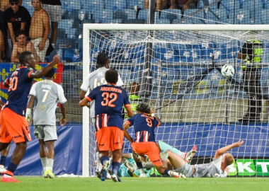 Soccer match scene: players in blue and orange chase the ball near the goal as the goalkeeper lies on the ground.