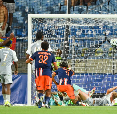 Soccer match scene: players in blue and orange chase the ball near the goal as the goalkeeper lies on the ground.