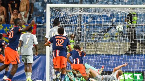 Soccer match scene: players in blue and orange chase the ball near the goal as the goalkeeper lies on the ground.
