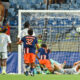 Soccer match scene: players in blue and orange chase the ball near the goal as the goalkeeper lies on the ground.