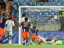 Soccer match scene: players in blue and orange chase the ball near the goal as the goalkeeper lies on the ground.