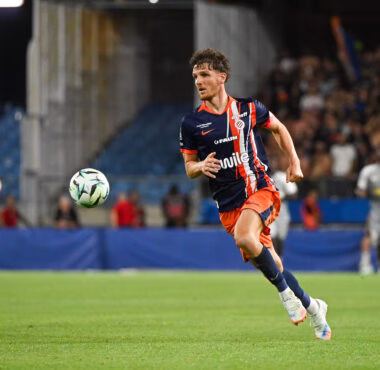 Soccer player in navy and orange kit runs toward the ball on a grass field with a blurred stadium crowd behind him.