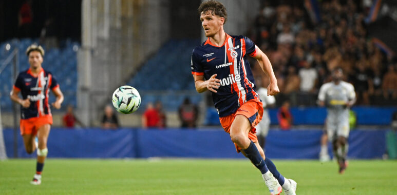 Soccer player in navy and orange kit runs toward the ball on a grass field with a blurred stadium crowd behind him.