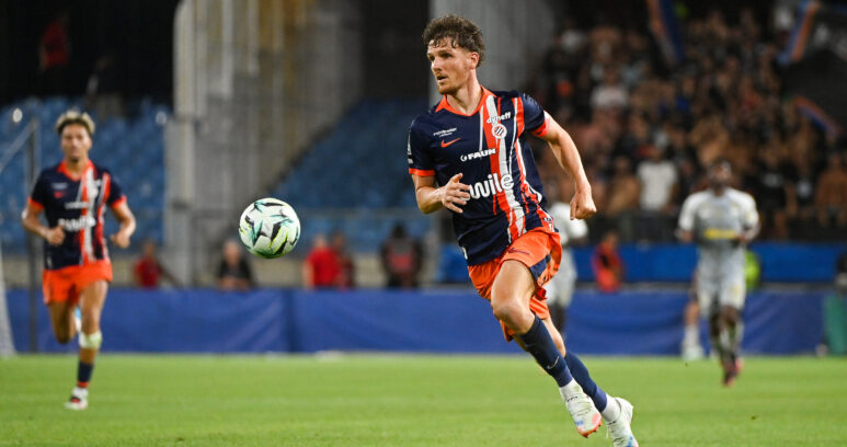 Soccer player in navy and orange kit runs toward the ball on a grass field with a blurred stadium crowd behind him.