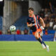 Soccer player in navy and orange kit runs toward the ball on a grass field with a blurred stadium crowd behind him.