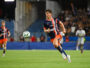 Soccer player in navy and orange kit runs toward the ball on a grass field with a blurred stadium crowd behind him.